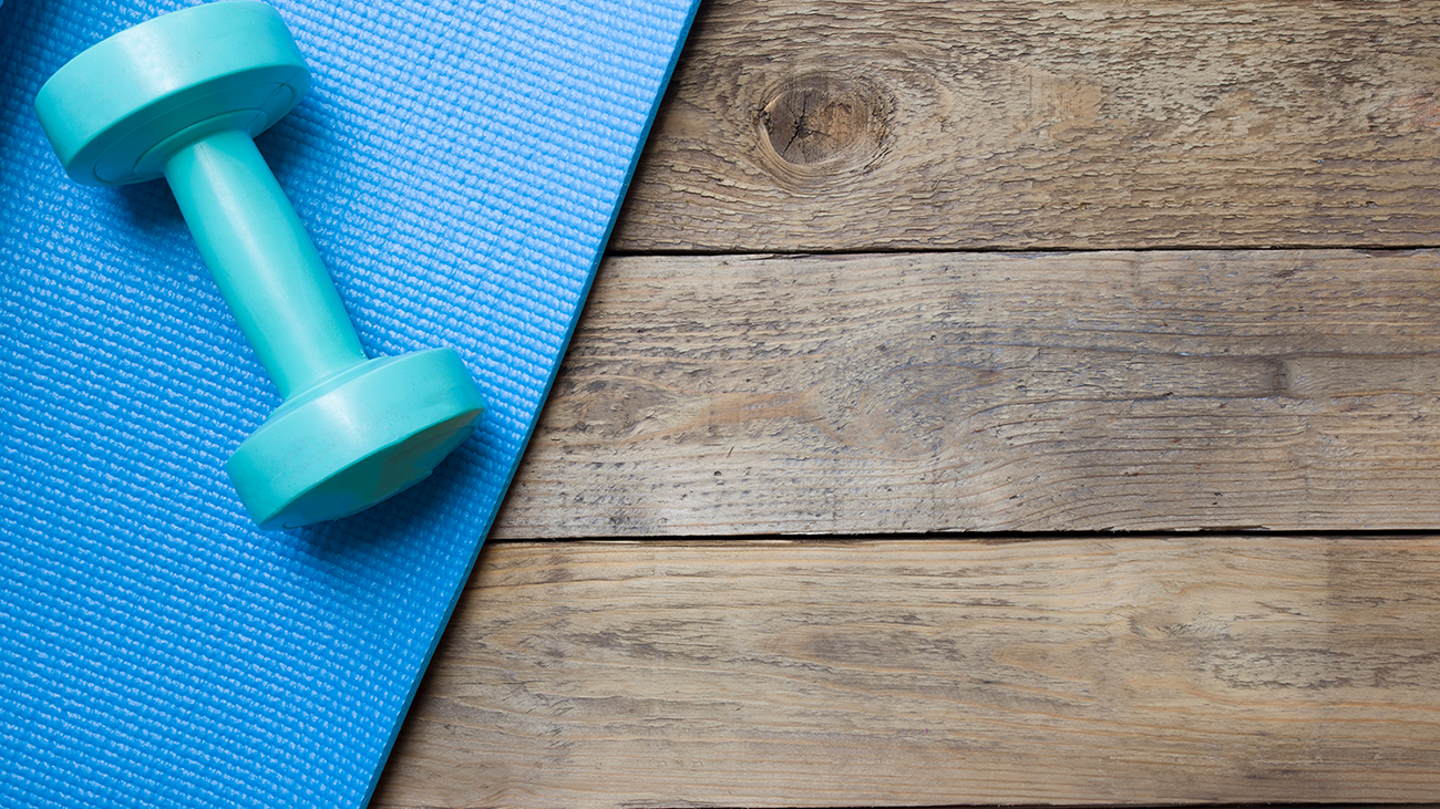 Image of an exercise mat laying on a wood floor. On the exercise mat is a dumbbell.