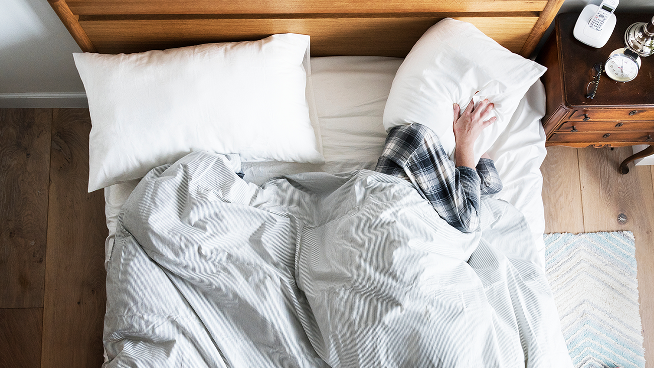 Image of someone laying in their bed with a pillow wrapped around their head. Their hands are holding the pillow in place over their ears to muffle any noise.