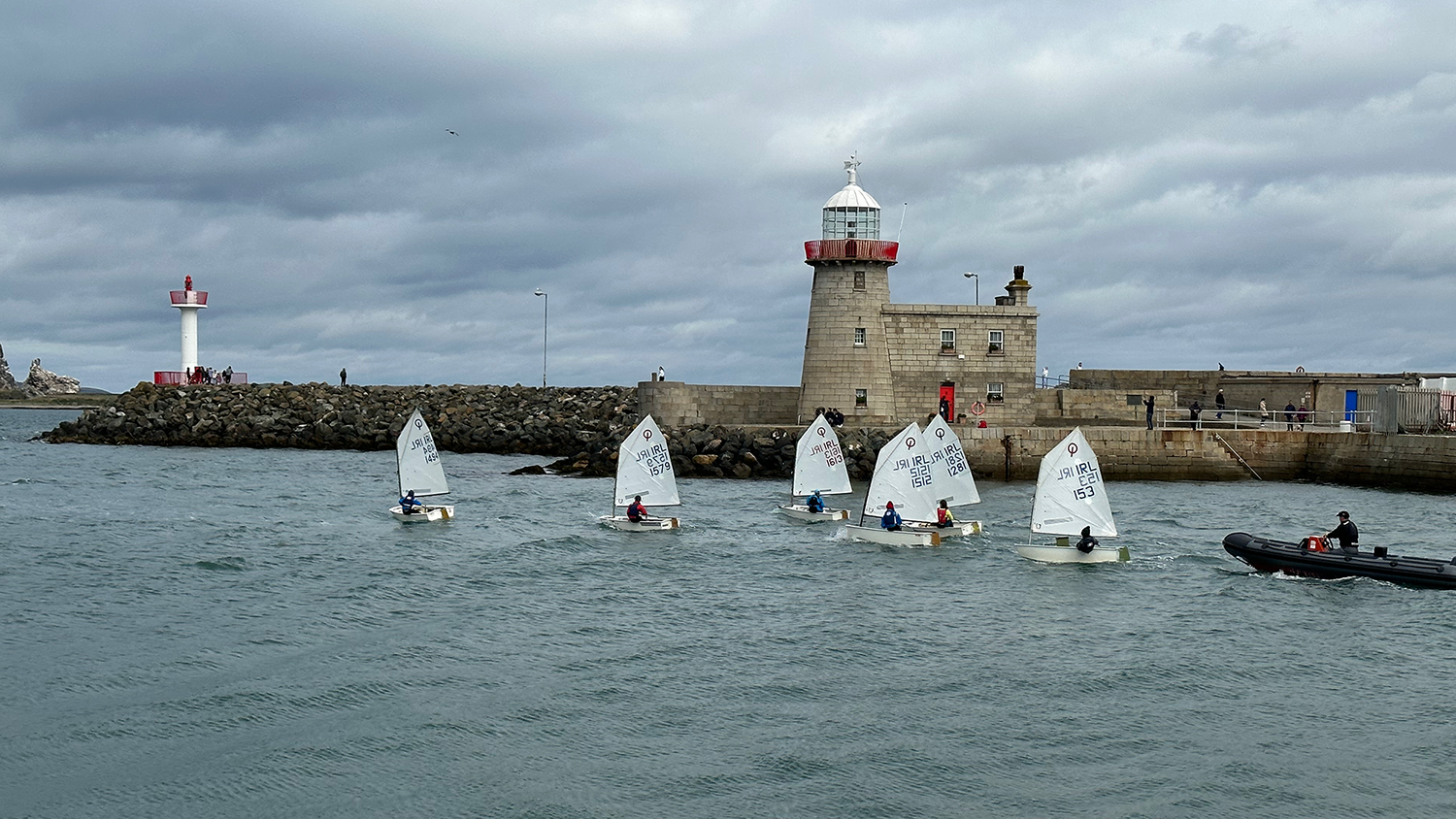 Children in individual sailboats taking sailing lessons with their instructors. In the background of the photo, you can see two lighthouses along the coast of Howth, Ireland.