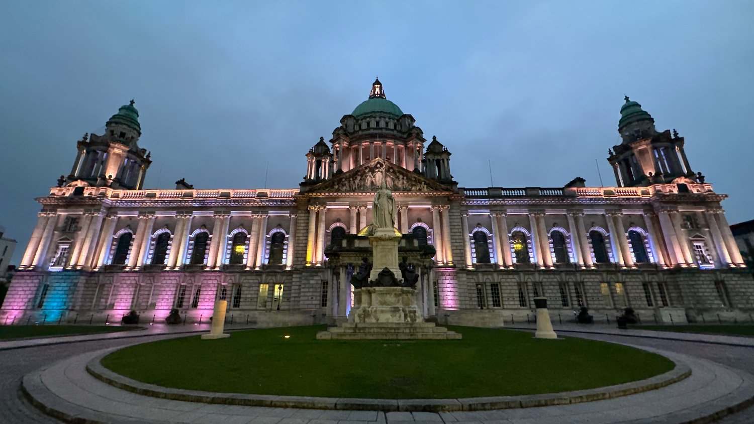 A photo of the Belfast City Hall at night. The building has dramatic lighting illuminating the building's stone columns and statues.