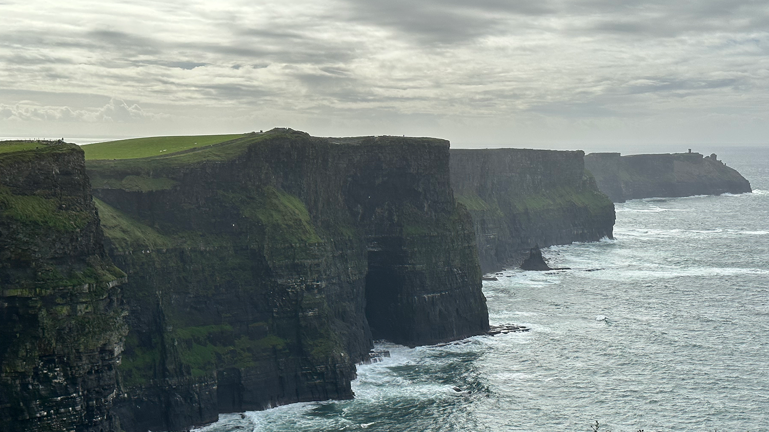 A photo with the Cliffs of Moher. Waves are crashing into the cliffs. There are spotty clouds in the sky with the sun peaking through.